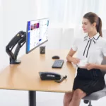 אישה יושבת ליד שולחן עם שולחן שולחנית דו מפרקית ארגונומית ל2 מסכי מחשב עד "27 וכוס קפה.