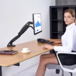 אישה יושבת ליד שולחן משרדי ועובדת על מחשב עם זרוע ארגונומית כפולת מפרקים למסך 30 אינץ'.