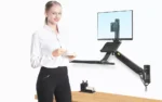 אישה עומדת מחייכת כשהיא מחזיקה ספל וצלוחית ליד עמדת עבודה ארגונומית מיועדת לתלייה על קיר למסך מחשב, מקלדת ועכבר מעל שולחן עבודה עם ציוד משרדי.