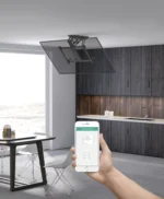 יד אוחזת סמארטפון עם ממשק אפליקציית בית חכם במטבח מודרני עם קולט אדים מתקפל חשמלי תקרה זרוע חכם למסכים עד 70 אינץ' מעל.