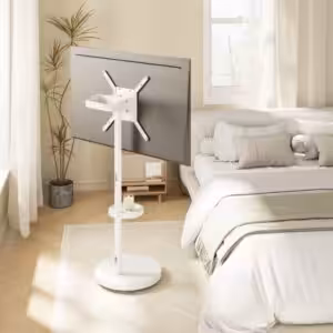 חדר שינה מודרני עם טלוויזיה המותקנת על מעמד רצפתי לבן ליד המיטה, עם מצעים ניטרליים ועציץ בפינה. המעמד מתאים למסכים בגודל של עד 65 אינץ' ומציע סגנון ושימושיות רב-תכליתיים.